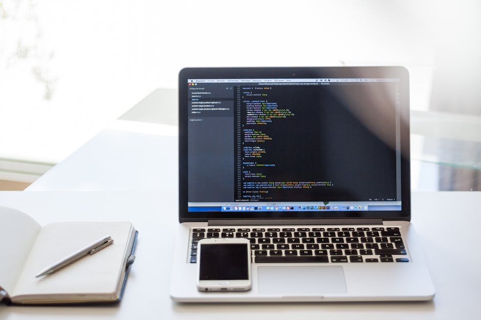 The image shows a modern office desk with a silver laptop displaying lines of code on a dark background, positioned centrally. To the left of the laptop is an open white notebook with a silver pen res